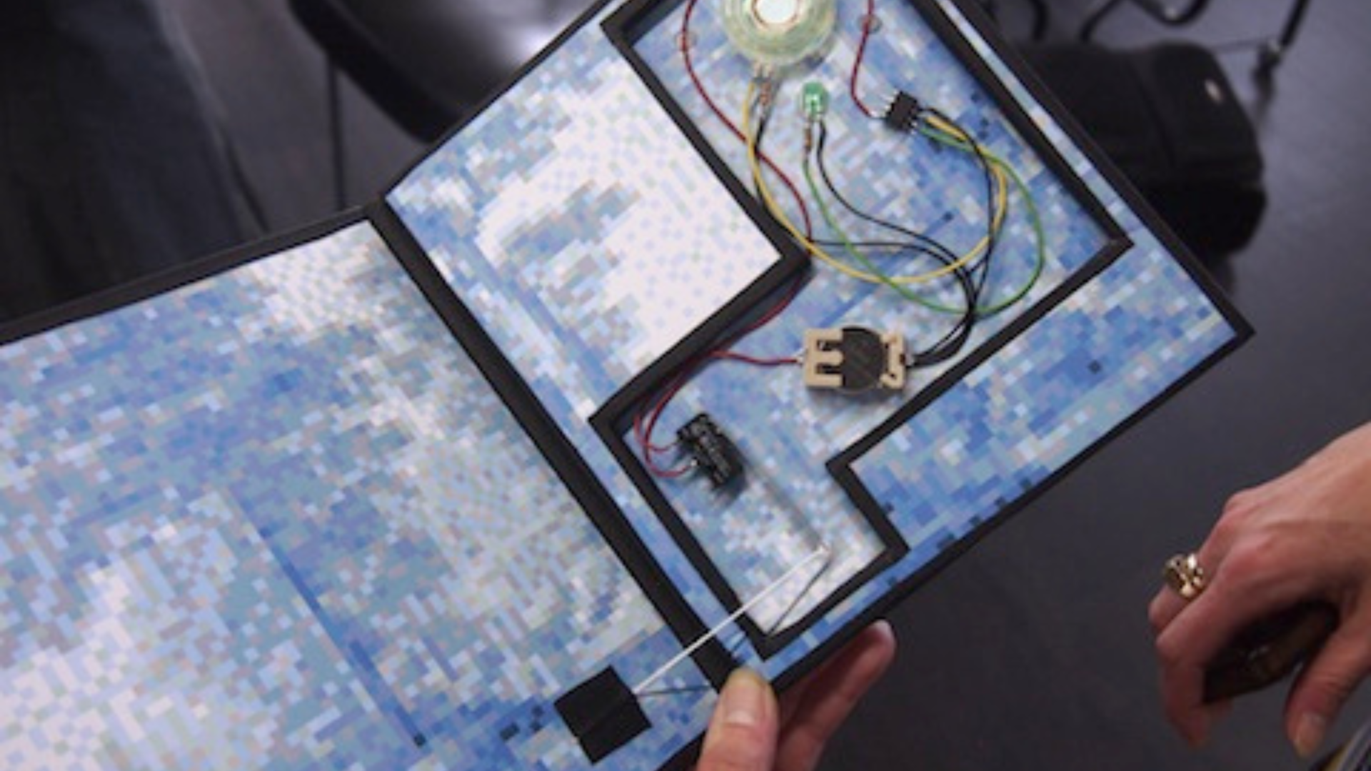 Close-up of hands holding an open book with digital components. The page is pixelated blue, featuring wires and a light, suggesting interactive technology.