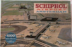 Illustrated puzzle box cover of Schiphol Airport, Amsterdam. Shows an aerial view with planes at terminals, emphasizing scale and complexity. Text highlights "over 1000 pieces."