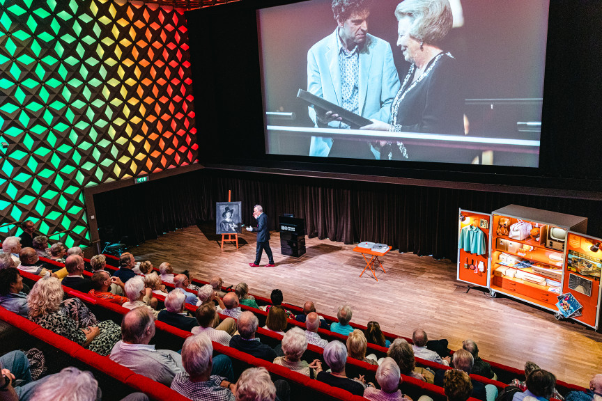 Een zaal met senioren kijkt naar het televisienostalgie arrangement. Ze zien het fragment waar Koningin Beatrix het gebouw van Beeld & Geluid opent.