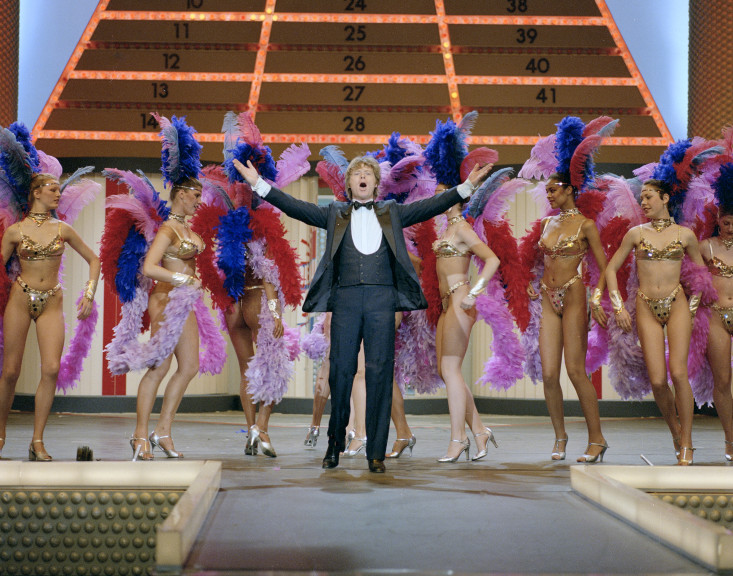 Willem Duys staat op het podium met showgirls op de achtergrond