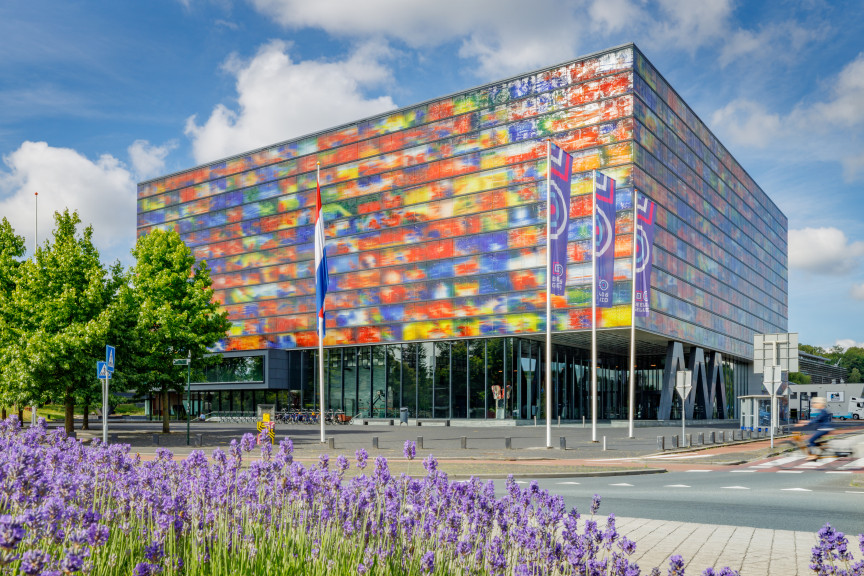 Het kleurrijke vierkant gebouw van Beeld & Geluid voor een blauwe, licht bewolkte lucht. Op de voorgrond ontpoppen zich paarse bloemen tijdens een lentedag