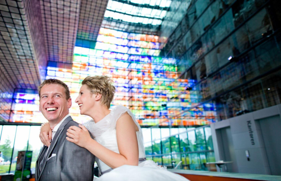 Een blij stel in trouwkledij (grijs pak en witte jurk) in het atrium van Beeld & Geluid. Op de achtergrond zie je de gekleurde glazen ramen