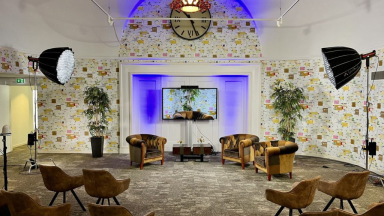 Zaal in Beeld & Geluid Den Haag voor de webinar: setting met drie sprekersstoelen en in het midden een tafel, achter aan de muur een tvscherm. In de zaal zie je verder een stoelopstelling gericht op de sprekersstoelen en aan beide kanten grote lampen om de setting uit te lichten.